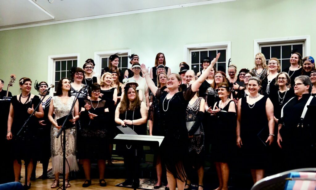 A group of women in black and white 1920's dresses have just finished singing. The chorus directer is standing in a victorious pose with her hands raised.