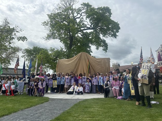 Many people, mostly women, sit or stand in front of a large object that is covered in fabric.