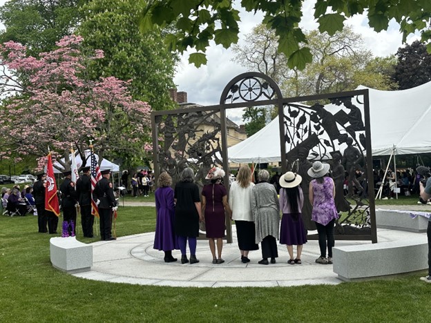 Several women stand in front of an imposing sculpture. It looks like a large gateway with a door. There are numerous carvings of women integrated into the gate.