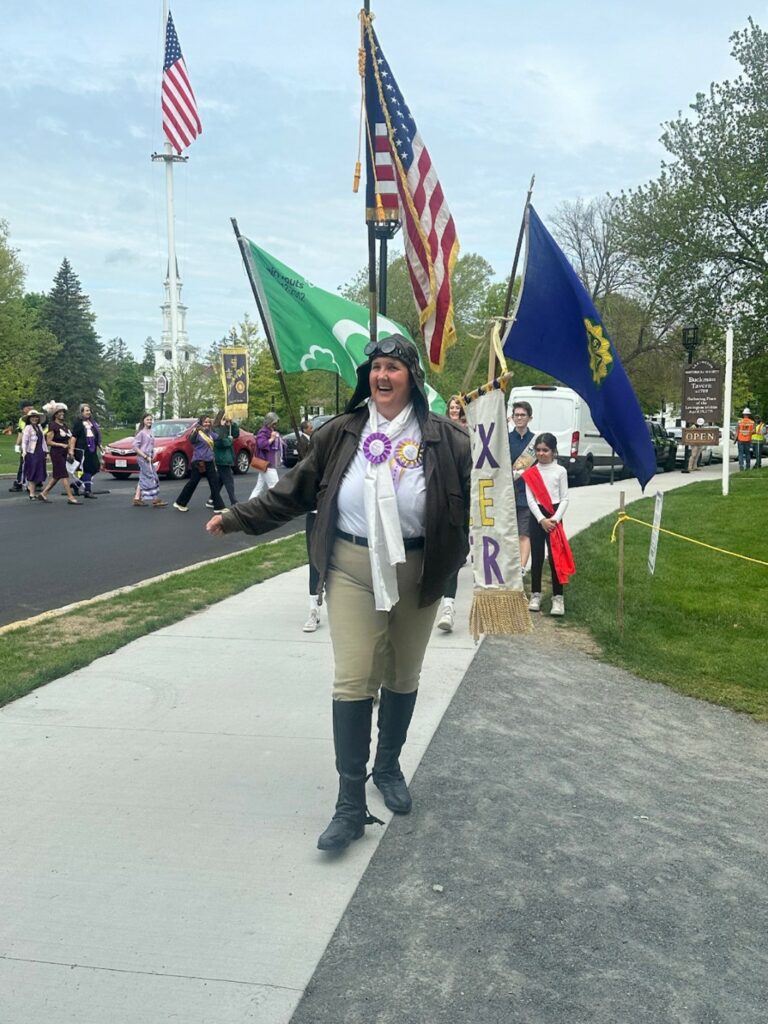 A woman wearing 1920's aviator gear -- a leather aviator hat and jacket is walking. Several flags are right behind her, and other people walking, parade-style, are in the background.