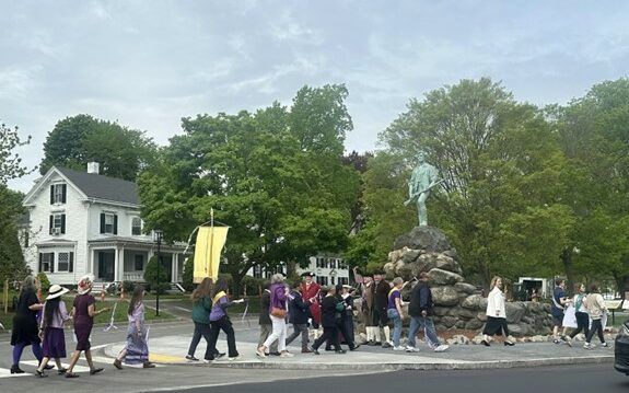 A number of people walking on a street. Lexington's Minuteman statue is in the background.