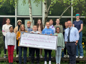 A group of 7th grade students present an oversize check for the Acton Food Pantry to a smiling woman.