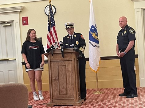 A woman in a formal fire-fighters uniform (complete with white hat) is speaking at the podium. A young woman in a t-shirt and tennis shoes stands on one side, and a man wearing a police uniform stands on the right.