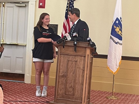 A man wearing a suit and tie stands at a podium. He is presenting an award to a young woman dressed in a t-shirt and sneakers.