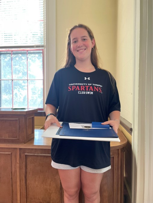A young smiling woman holds a stack of awards that she just won. She is wearing a University of Tampa Swim Club t-shirt.