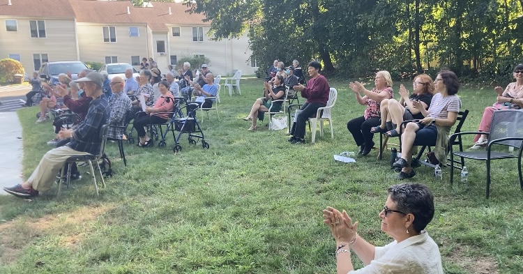 A group of people clapping. People are sitting in lawn chairs on the grass.