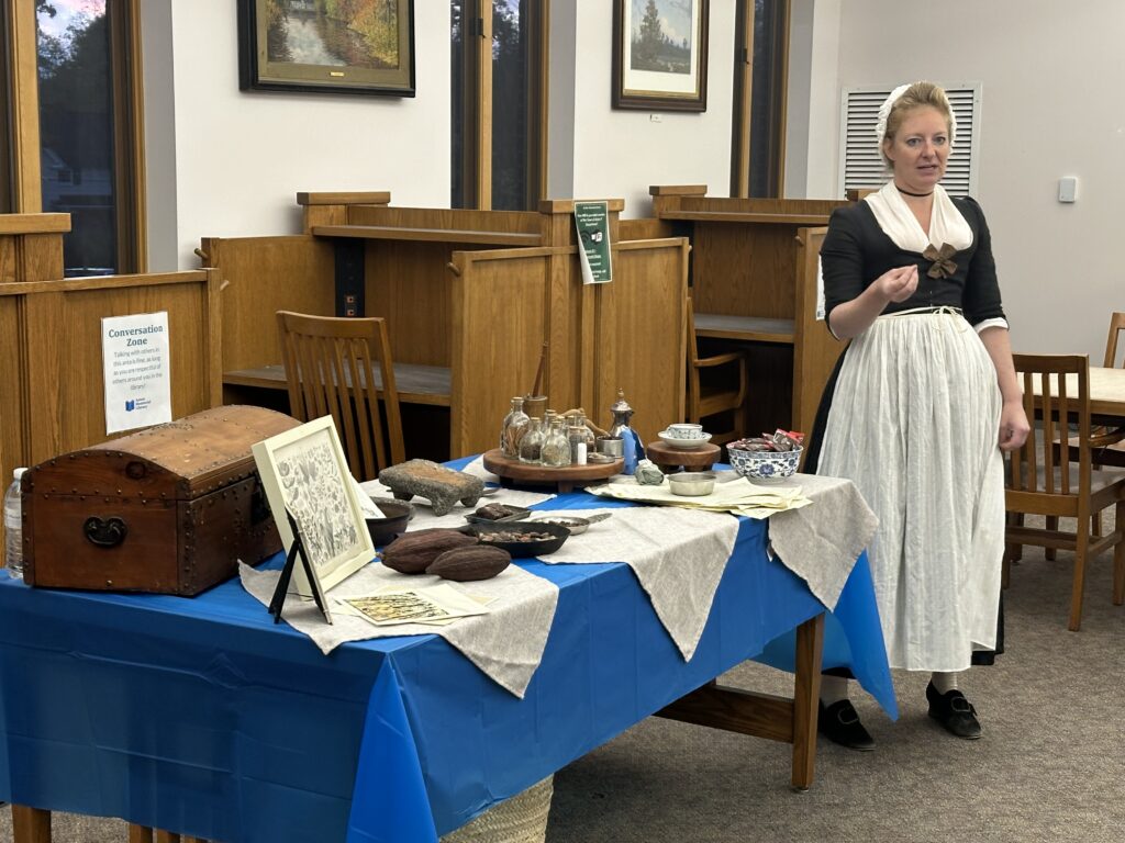 A woman wearing a colonial-style dress stands in front of a table covered with spices in little glass bottles, cacao pods, and other items related to 18th century chocolate manufacturing. A bowl of old-style chocolate samples is also available.