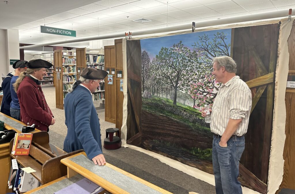 A man stands next to a mural that shows a field with fruit trees in bloom in the background. Several men wearing colonial garb admire the mural and chat with the artist.