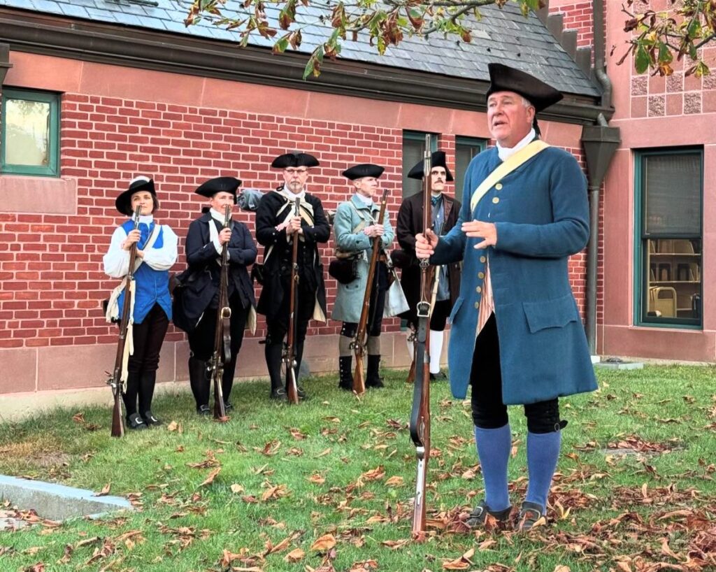 A man wearing a tricorner hat and blue colonial-style jacket stands by the library holding a musket. A group of men and women, also holding muskets (and wearing colonial clothing), stand in the background/