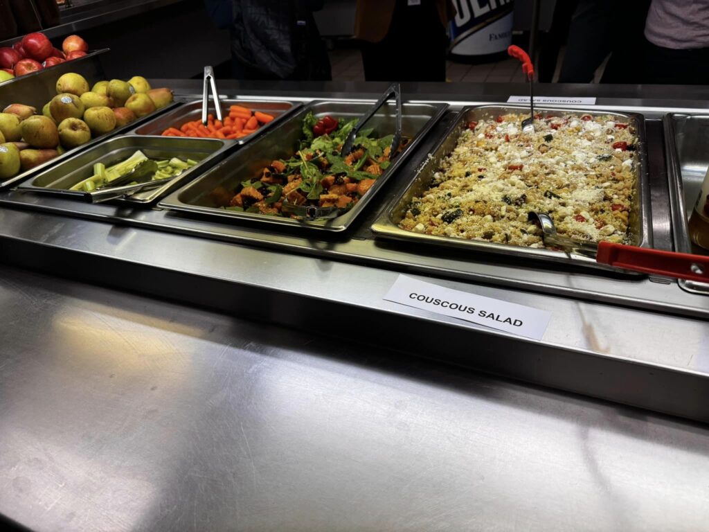 A cafeteria line-up, from left to right: 2 kinds of fresh apples; two kindsof raw veggies; a salad with spinach, roasted squash, and cranberries; and a couscous salad with lots of vegetables.