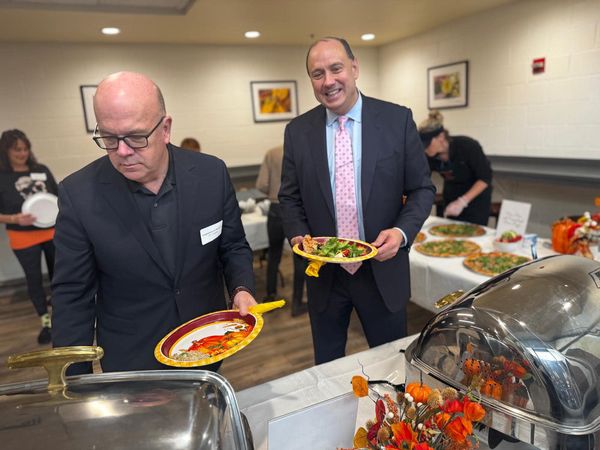 Two men in suits (one with a pink tie) stand in front of some steam trays with paper plates in hand.