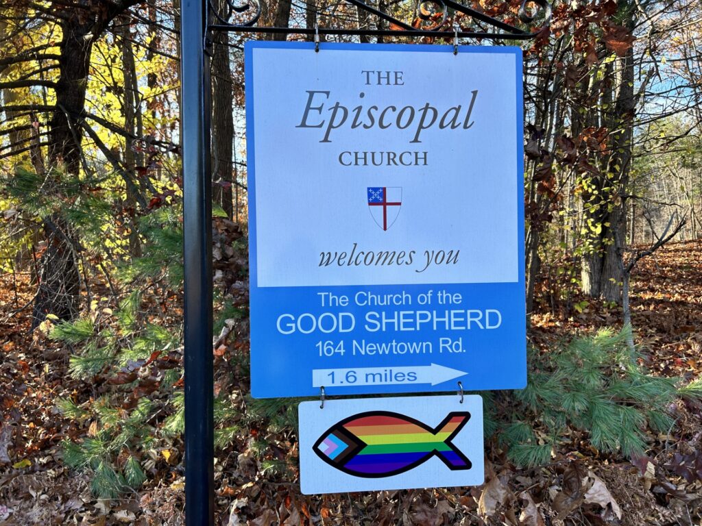 A sign to the Episcopal Church in Acton. A fish painted to represent all ofthe LGBTQ+ colors hangs from the bottom of the sign.