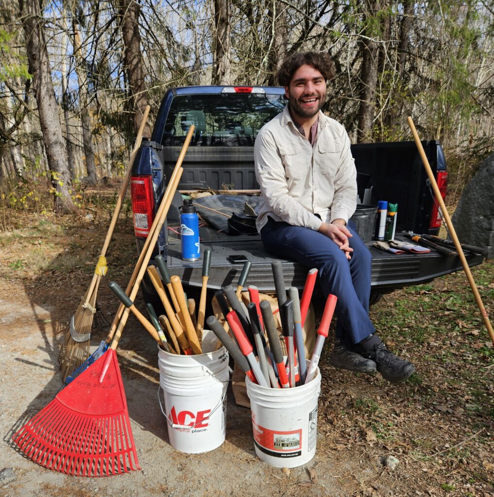 A smiling man sits on the edge of a truck with the back down.A couple of buckets of yard tools and some rakes are ready and waiting for the volunteers.