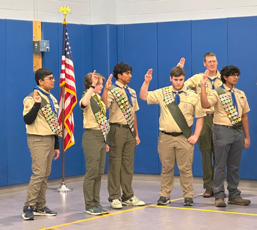 Five kids wearing scout uniforms (including well-decorated sashes) stand to take an oath in front of an American Flag. The scoutmaster is in the background.