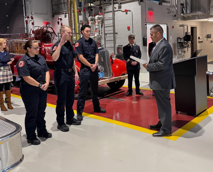 On the right, a man in a gray suit is reading from a piece of paper to three people dressed in firefighter uniforms. A firetruck is in the background and other people are watching.