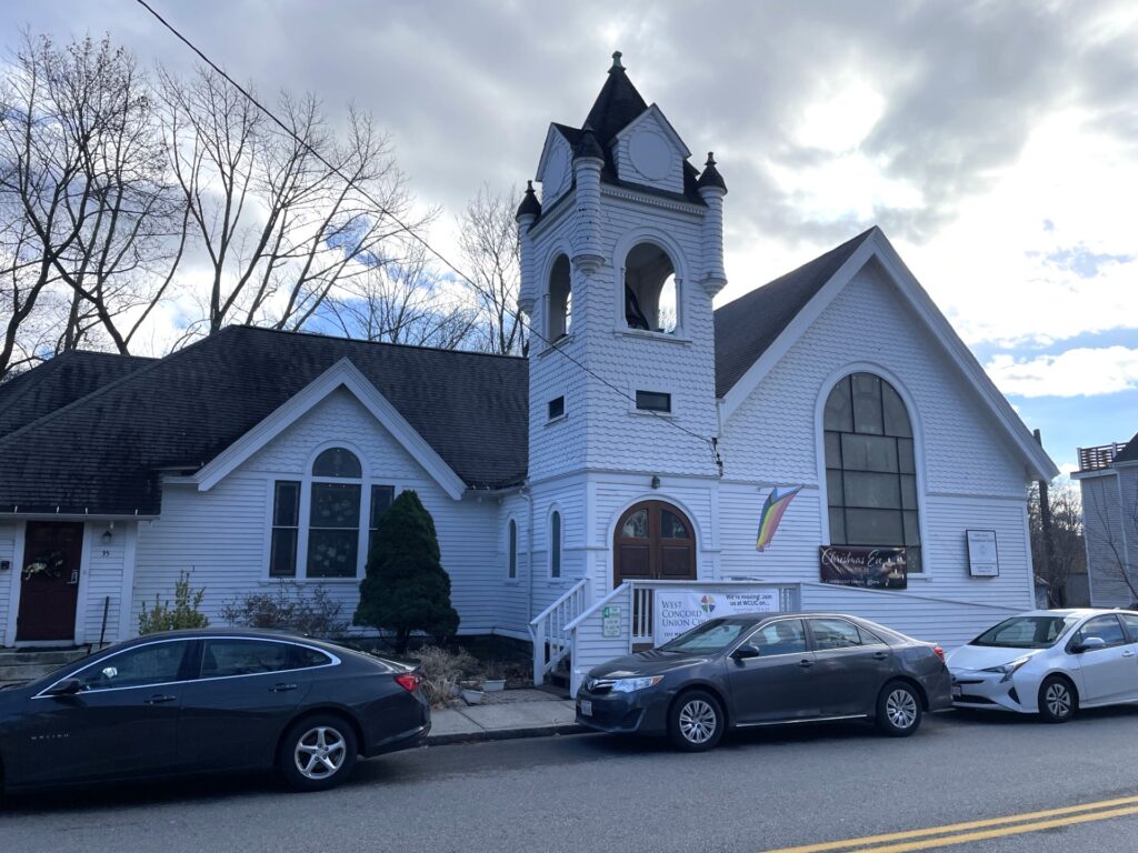 A large white church building right up against the street. The church flies a Pride flag.