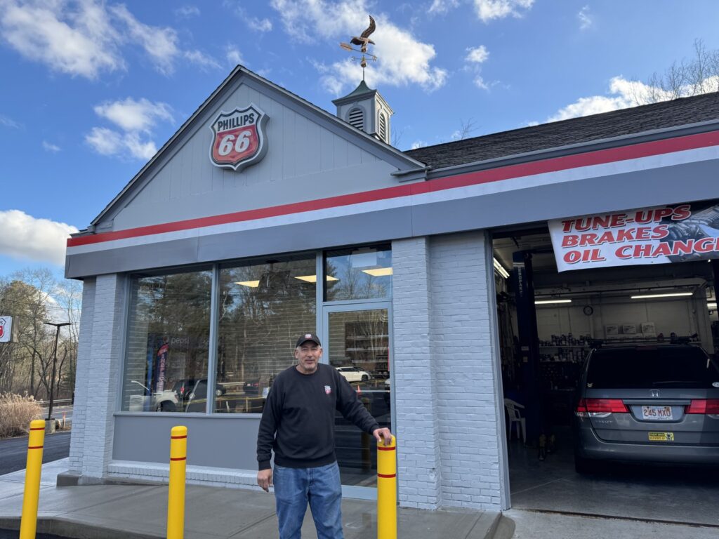 A man stands in front of a building with a Phillips 66 logo. There are yellow bollards in front of the building.