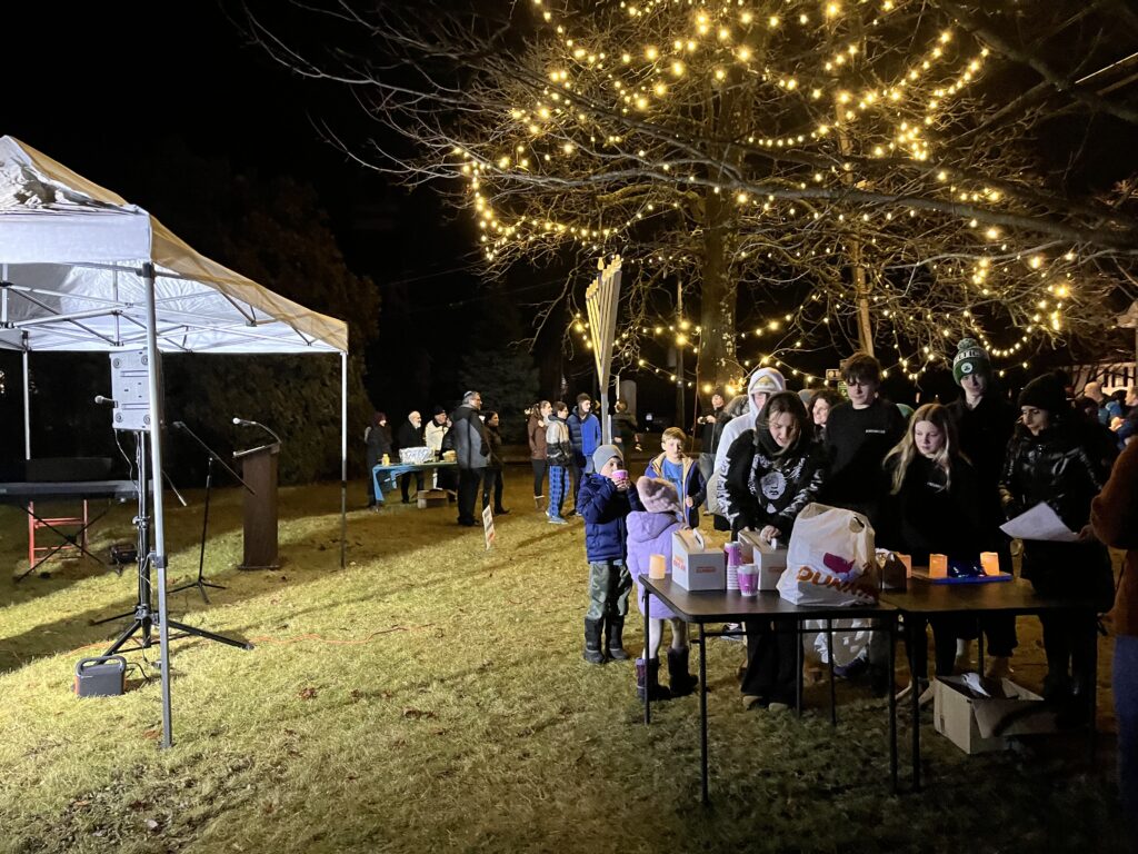 People are lined up in front of a table for donut holes and hot cocoa. It's dark out and a lit tree is in the background.