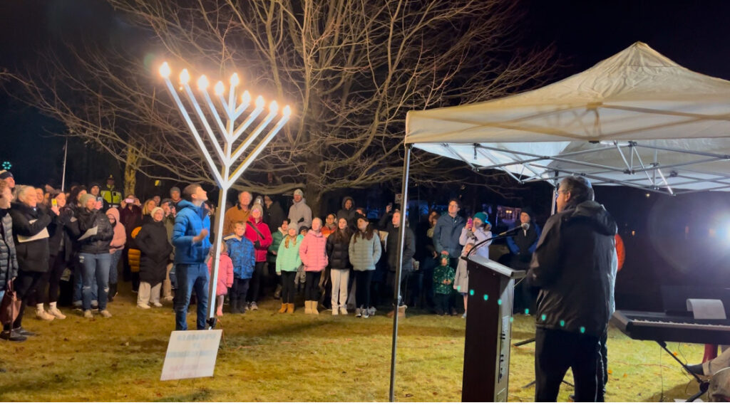 On the left, a 9-foot menorah is fully lit on a dark night. On the right, a man stands at a podium.