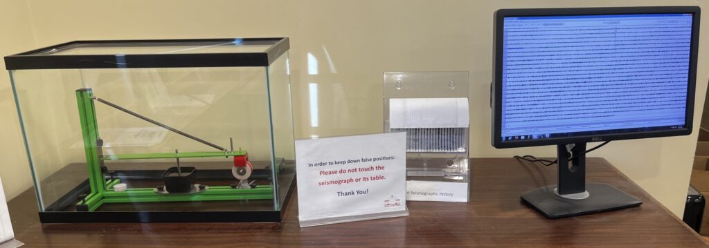 A table with a green seismometer in a fishtank (for extra protection, a seismograph printer, and a computer screen showing the printout in larger detail.