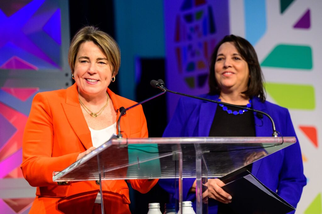 Two women, one with short hair wearing an orange jacket, the other with longer black hair wearing a blue jacket, stand in front of a podium.