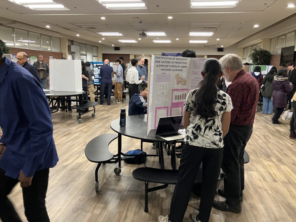 A student and an older man stand in front of a scientific poster. Behind them, you can see several other tables with similar posters.