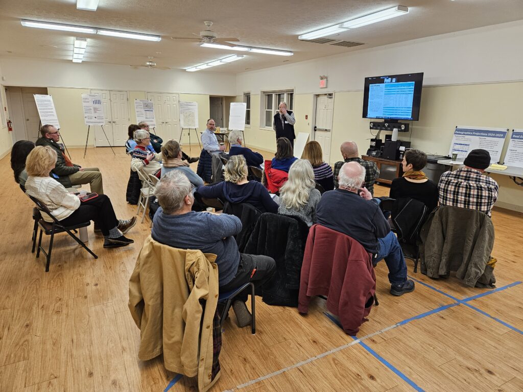 A group of people sitting in a room facing a large computer screen.