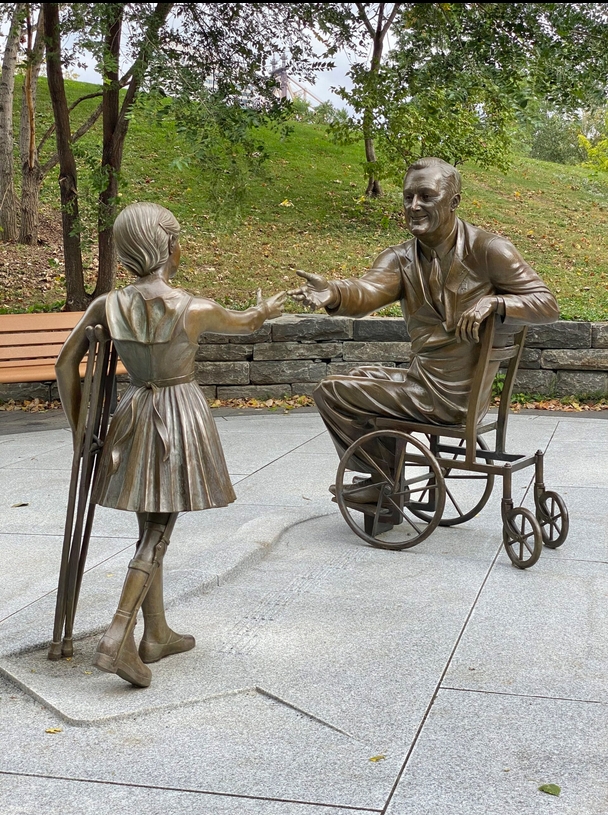 A bronze statue of a man in an old fashioned wheelchair reaching out to a girl (also a statue) who has a brace on her leg (from polio) and crutches.