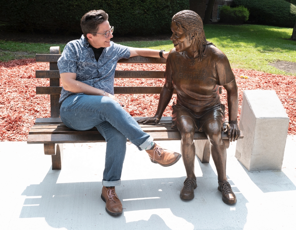 A bronze statue sits on a park bench in the sun. The smiling statue has hair in locks and is wearing an Ithaca is Gorges t-shirt. A person in a Hawaiian style shirt and jeans also sits on the park bench, They seem to be having a lively conversation.