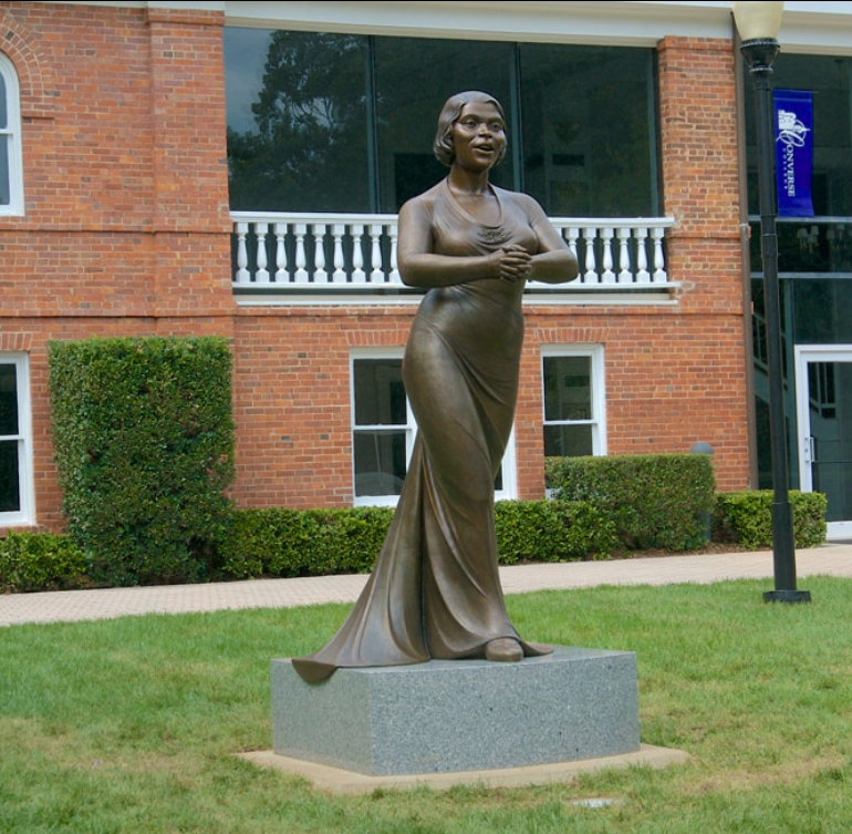 A statue of a woman in front of a brick building. The statue wears an evening gown. Her hands are clasped in front of her and she looks like she could be singing.