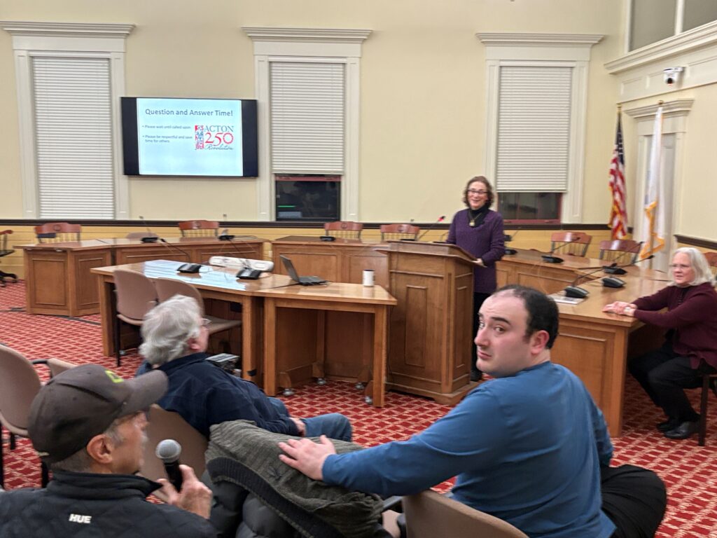 A woman stands at a podium. In the corner of a picture, a man in a baseball cap holds a mic. A few other people are in the picture.
