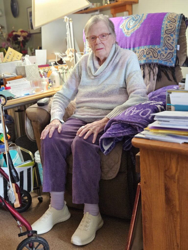 An older woman (she's 93!) sits in a comfortable chair. There's definitely a purple theme.