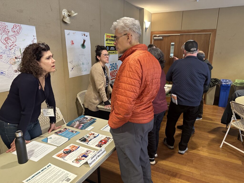 A woman leans on a table full of literature talking to a man with gray hair and an orange parka.