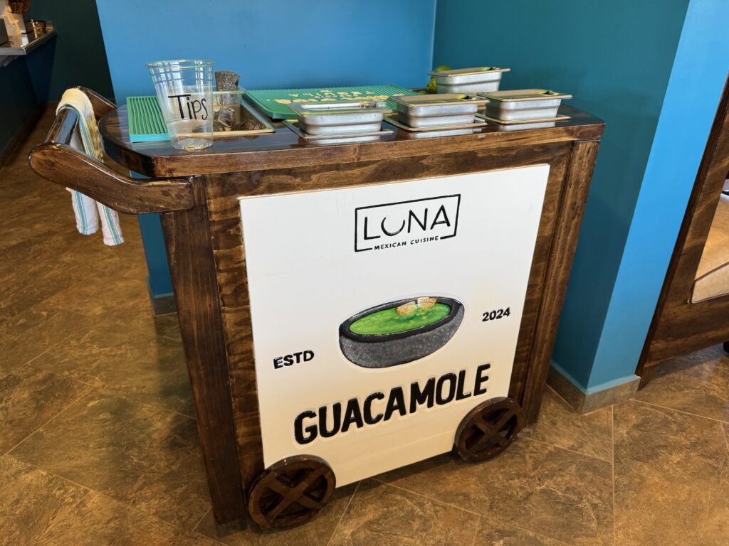 A wooden cart piled with condiment trays. There's a picture of a bowl of guacamole painted on the front of the cart and it's labeled Guacamole in large letters.