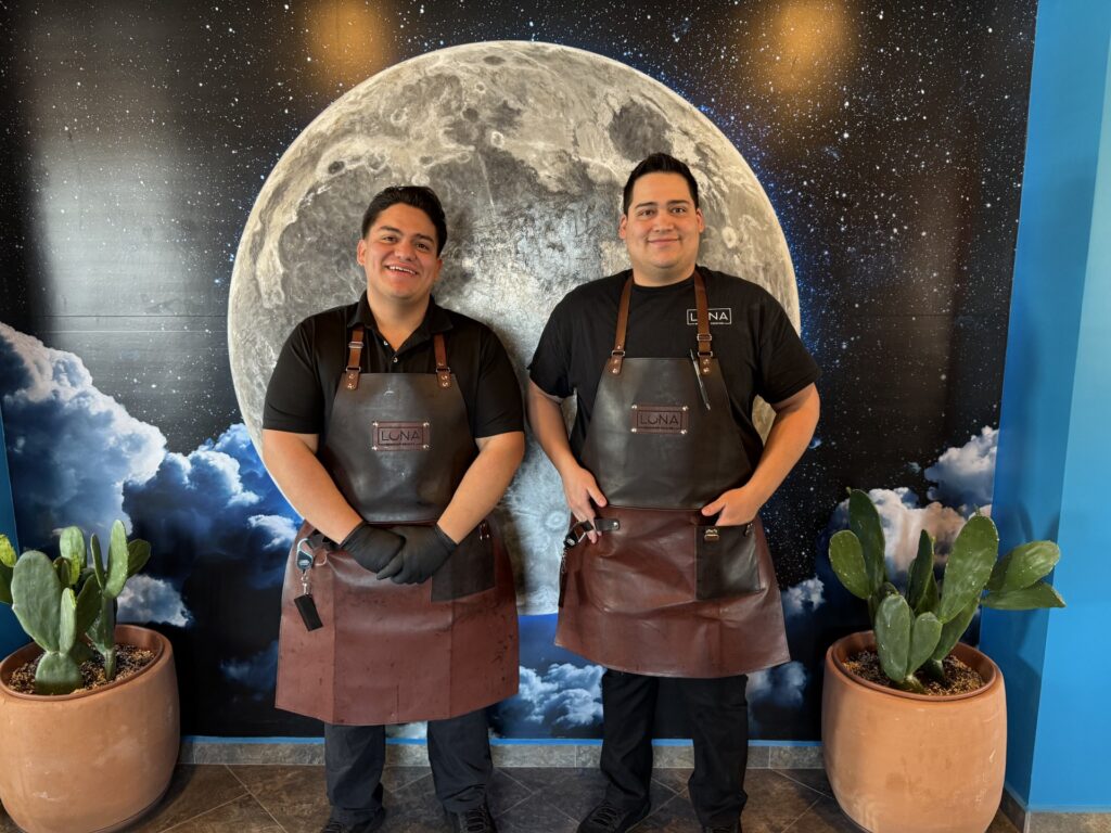 Two smiling youngish men wearing leather aprons stand in front of a painted moon. There are large cacti in clay pots on each side of the moon picture.