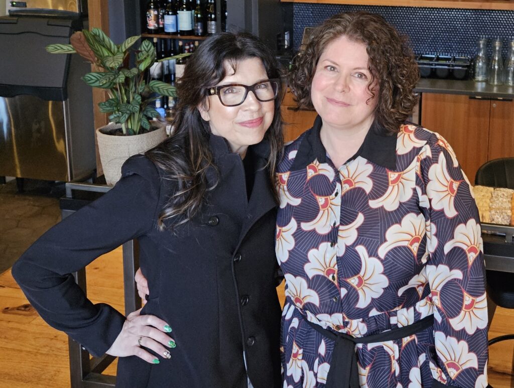 Two women, one with dark hair and glasses, the other with shorter, curlier hair, pose in their restauran, Silver Girl.