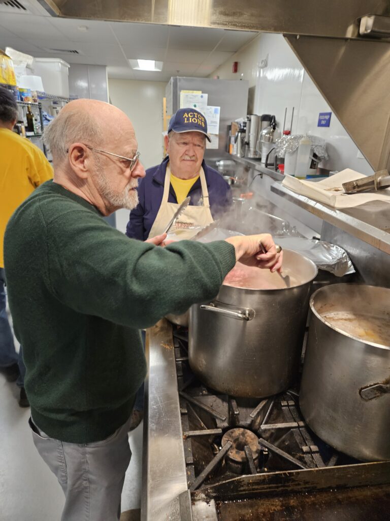 An older man wearing green stirs a large pot. Another man wearing an Acton Lions cap looks on.