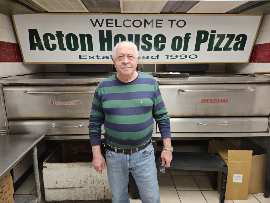 An older man in a striped shirt stands in front of a sign that says "Welcome to Acton House of Pizza, Established 1990"