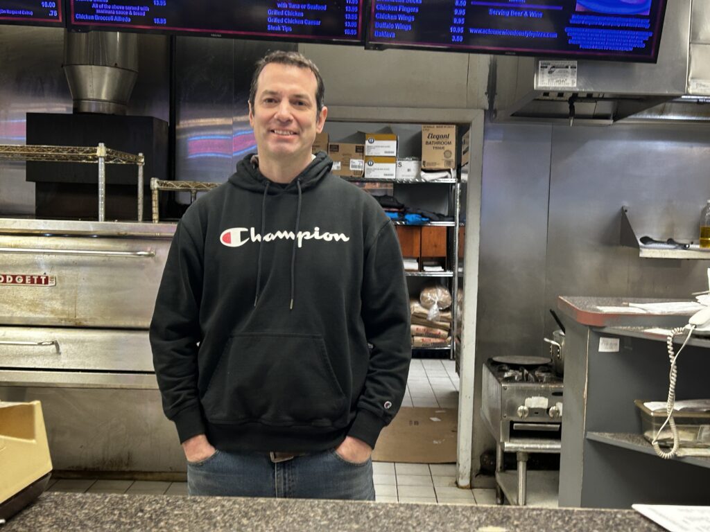 A man in a black sweatshirt stands at a counter with his hands in his pockets. A commercial kitchen is in the background.