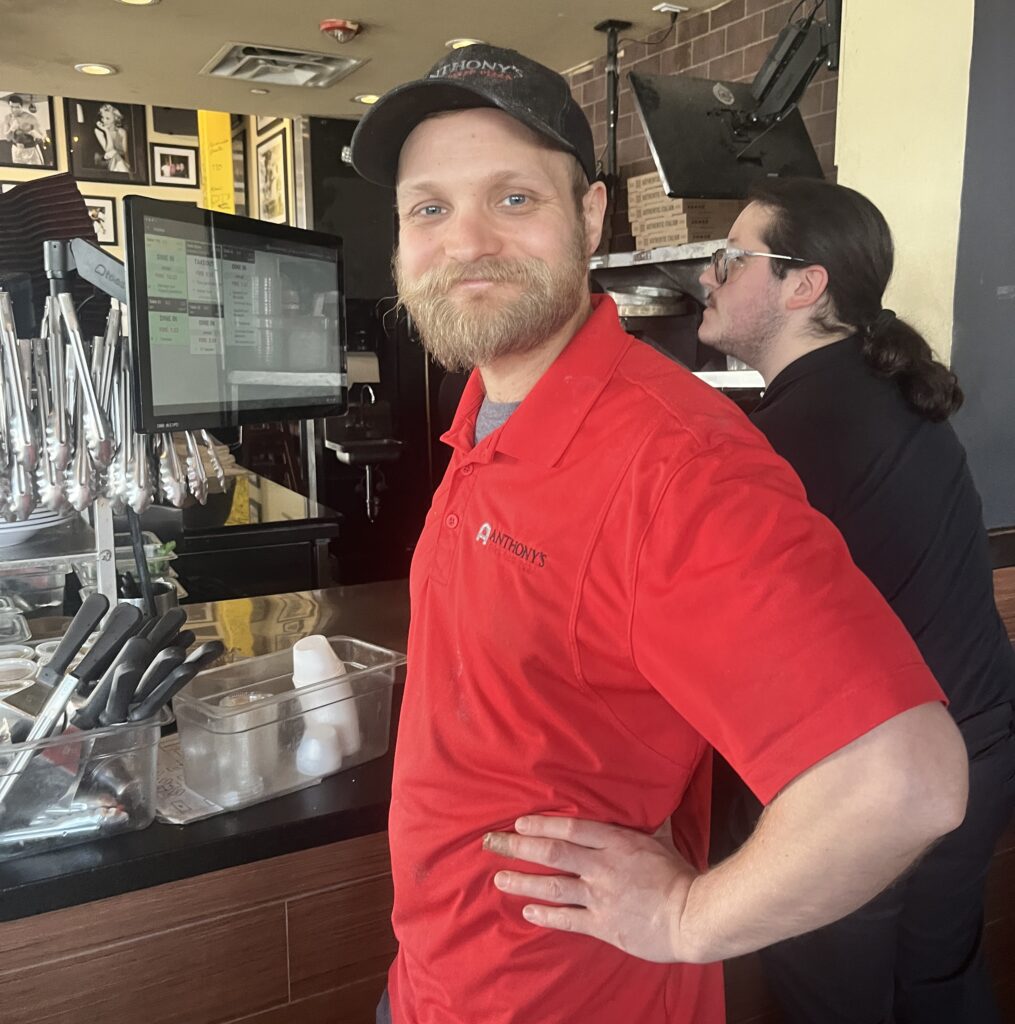 A bearded man in a red polo shirt stands in front of a commercial kitchen.