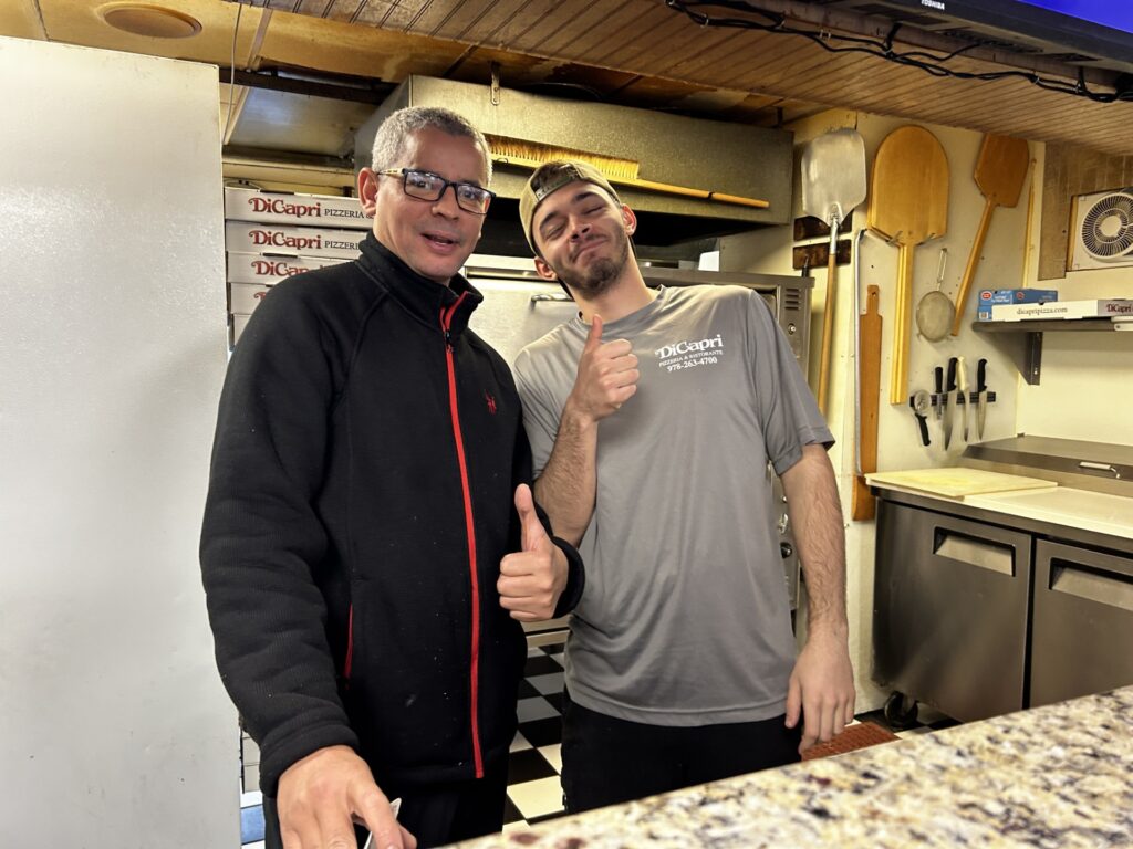 Two men, older and younger stand in a commercial kitchen. The younger man gives a big thumbs up.
