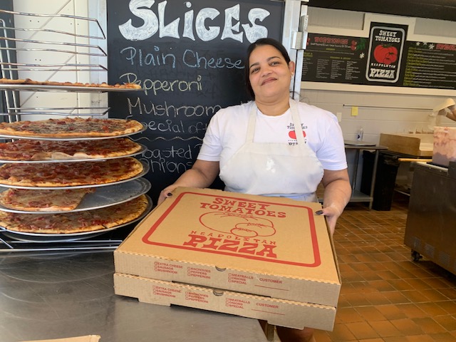 A young woman holds a couple of pizza boxes. A big stack of cooked pizzas on a rack is on her left. Yum!