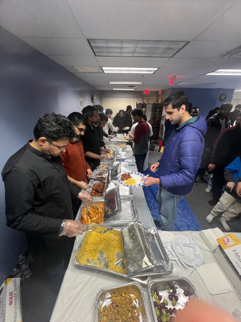 Men serve themselves from trays of food along a long table in a modern room with flourescent ceiling lights.