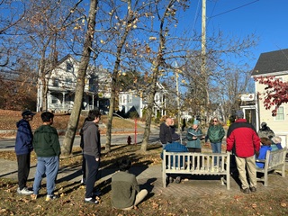 A group of people outdoors in front of a park bench. Local buildings are in the background.
