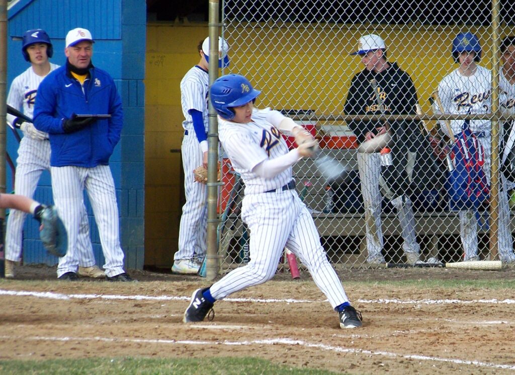 A player in a Revs uniform swings the bat. There are several people behind him including the catcher, the coach, and the next batter up.