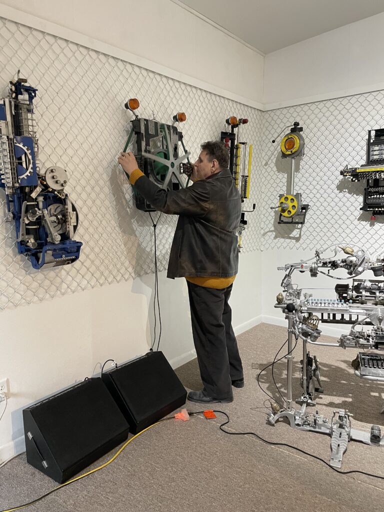 A man stands at a wall where several very interesting looking musical "instruments" are hanging. the instruments are mostly made from scrap metal. The man is standing in front of one instrument pulling on some straps, and the instrument is attached to a set of speakers.
