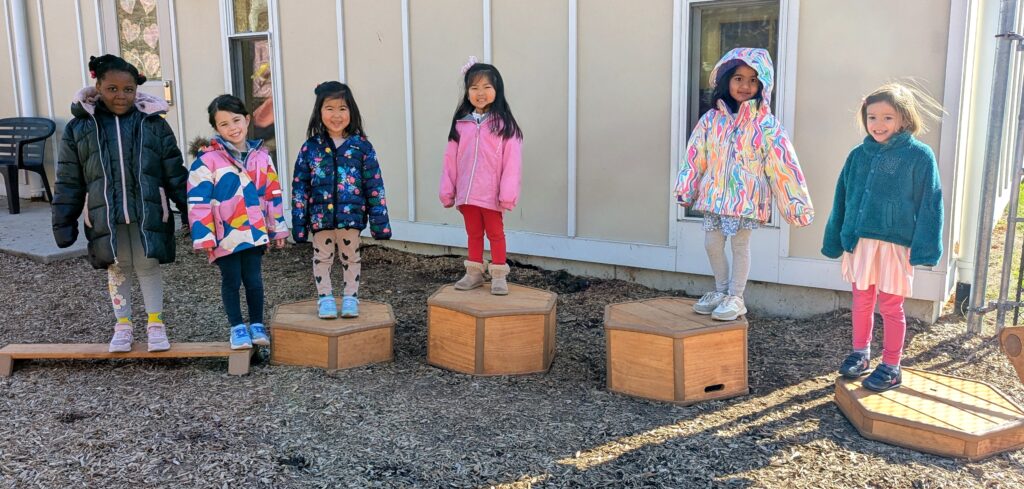 Six pre-school girls in colorful winter coats smile from their perches on mostly hexagonal, varying height, wooden play platforms in an outdoor, woodchip play yard, against a beige wall with white trip and two windows.
