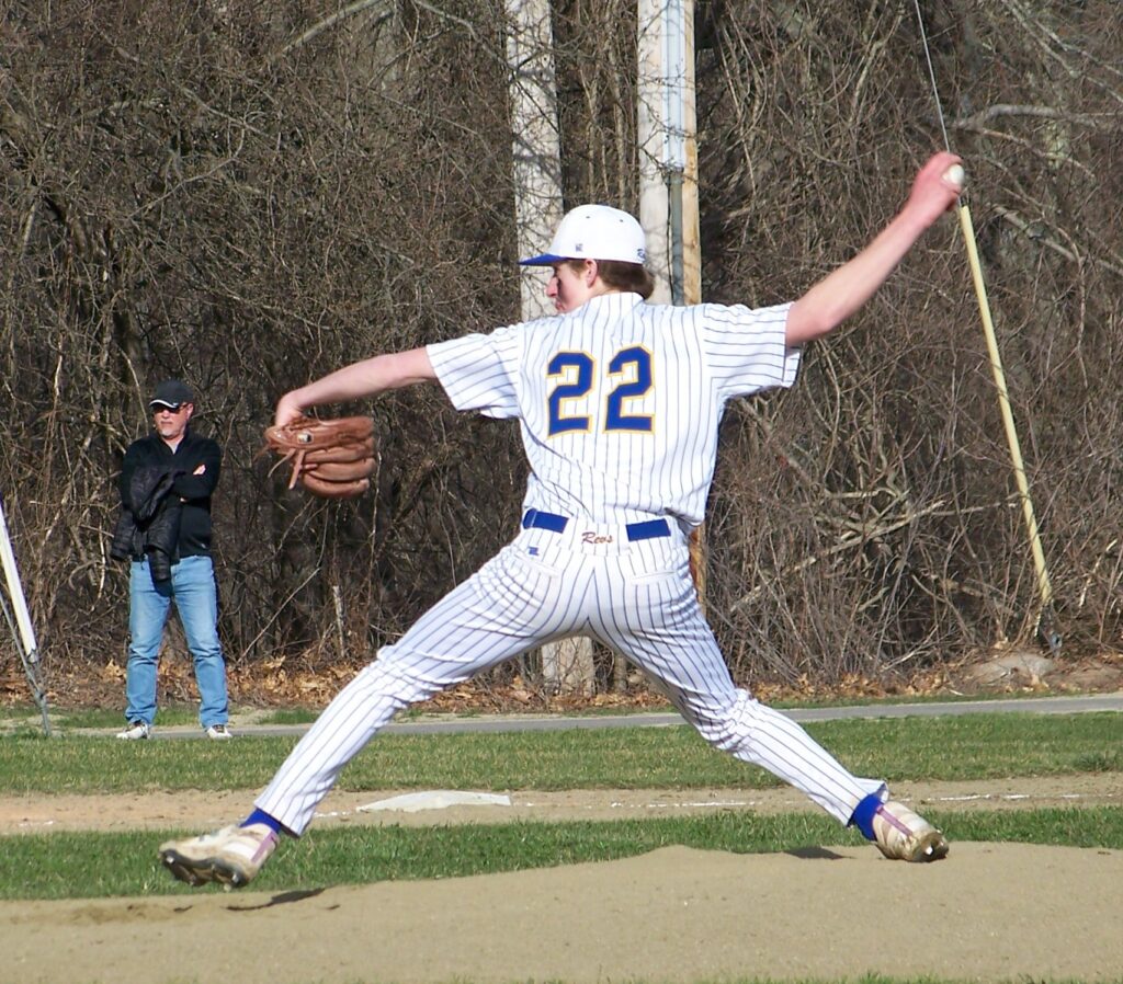 A baseball player in a Revs uniform is caught mid-pitch, with his arm behind his head.