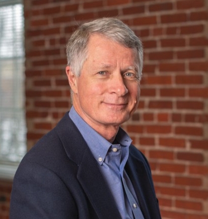 A grey-haired man in front of a brick wall.