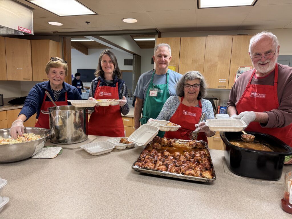 Five people in red aprons serve up baked beans, BBQ chicken, corn, and coleslaw.